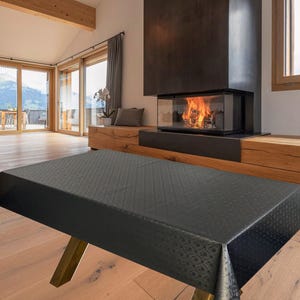 May include: A black patterned tablecloth on a wooden table with gold-toned legs. The table is in a modern living room with a fireplace and large windows. The room has wooden beams and flooring, with a view of the outdoors.