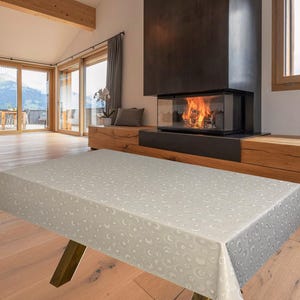 May include: A light grey rectangular tablecloth with a repeating crescent moon pattern. The tablecloth is draped over a wooden table with angled legs. The table is in a room with a fireplace and large windows.