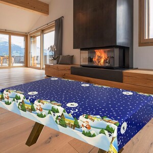 May include: A blue rectangular tablecloth with a winter scene border, featuring snow-covered houses, trees, and Santa Claus figures. The tablecloth has a pattern of white snowflakes and is on a wooden table.