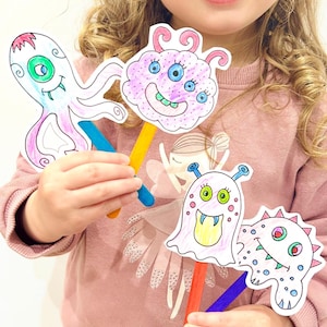 May include: Four colorful monster puppets on wooden sticks. The puppets are hand-drawn with various designs, including an octopus, a cloud-like creature, and two other unique monsters. The puppets are held by a person wearing a pink sweater.