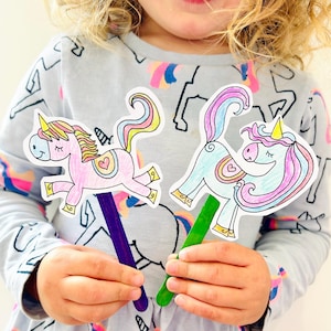 May include: Two colorful unicorn stick puppets. One is pink with a purple stick, and the other is blue with a green stick. Both have rainbow manes and tails, and are held by a person wearing a gray unicorn-themed shirt.