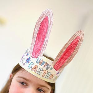 Puede incluir: Una diadema de orejas de conejo de Pascua hecha a mano. Las orejas son marrones con interiores rosas. La diadema es blanca con las palabras "Hoppy Easter" y diseños florales coloridos.