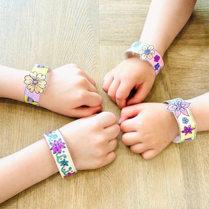 May include: Four paper bracelets, each adorned with colorful floral designs, are displayed. The bracelets, featuring various flower patterns in shades of purple, pink, yellow, and blue, are worn on wrists. The background is a light brown wooden surface.