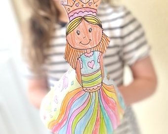 May include: A colorful, hand-drawn princess paper doll with a crown, long orange hair, and a rainbow-striped dress. The doll is held up in front of a blurred background. The princess's dress has a heart on the bodice.