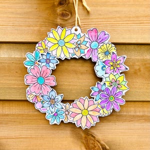 May include: A colorful floral wreath with a variety of hand-drawn flowers in shades of pink, yellow, blue, and purple. The wreath is made of paper and has a string for hanging. The background is a wooden surface.