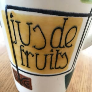 May include: A white ceramic jug with a yellow, black and green geometric design. The jug has the words "jus de fruits" written in black on the yellow section.
