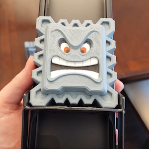 May include: A gray, 3D-printed Thwomp character from the Super Mario video game series. The Thwomp has an angry expression with orange eyes and a wide, toothy grin. It is mounted on a black metal frame.