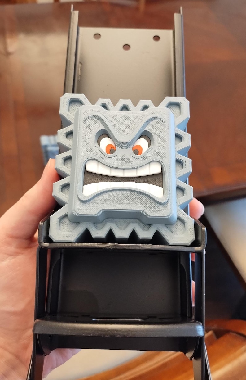 May include: A gray, 3D-printed Thwomp character from the Super Mario video game series. The Thwomp has an angry expression with orange eyes and a wide, toothy grin. It is mounted on a black metal frame.