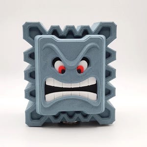 May include: A gray, 3D-printed Thwomp from the Super Mario video game series. The block-shaped character has a menacing expression with red eyes, a wide open mouth, and jagged edges.