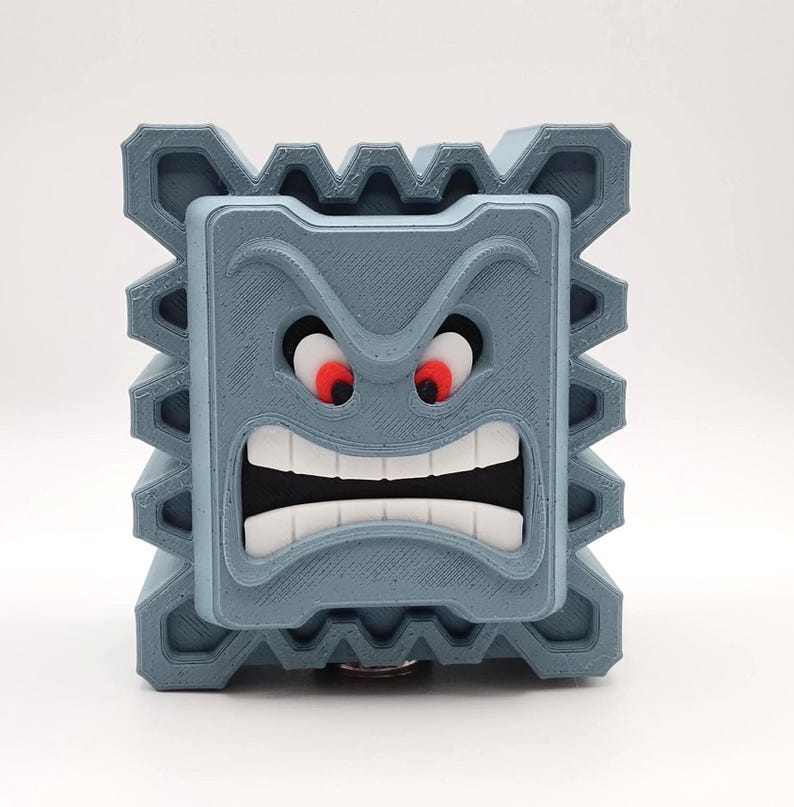 May include: A gray, 3D-printed Thwomp from the Super Mario video game series. The block-shaped character has a menacing expression with red eyes, a wide open mouth, and jagged edges.