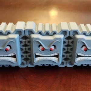 May include: A 3D-printed Thwomp from the Super Mario video game series. The gray, block-shaped character has an angry face with red eyes and a wide, toothy grin. The top of the block is white and ridged.