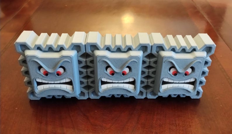 May include: A 3D-printed Thwomp from the Super Mario video game series. The gray, block-shaped character has an angry face with red eyes and a wide, toothy grin. The top of the block is white and ridged.