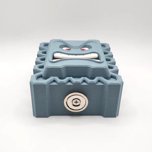 May include: A blue-gray, 3D-printed box with a cartoonish face on top. The face has red eyes, white teeth, and an angry expression. A silver lock is on the front of the box. The box has a textured, geometric design.