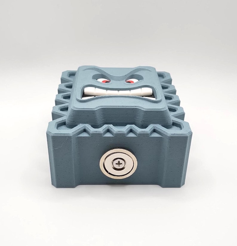 May include: A blue-gray, 3D-printed box with a cartoonish face on top. The face has red eyes, white teeth, and an angry expression. A silver lock is on the front of the box. The box has a textured, geometric design.