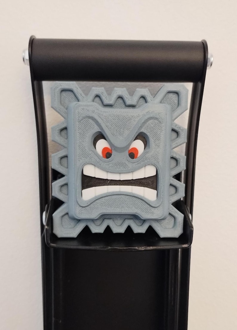 May include: A gray, 3D-printed Thwomp character from the Super Mario video game series. The Thwomp has an angry expression with red eyes and a wide open mouth. It is mounted on a black metal bracket.