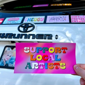 May include: A pink and white sticker with a rainbow gradient background. The sticker says "SUPPORT LOCAL ARTISTS" in colorful, stylized letters.