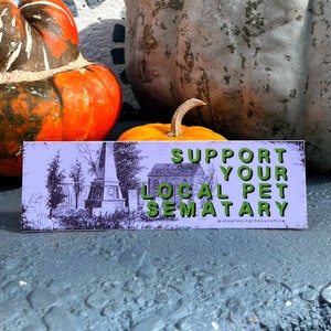 May include: A rectangular sign with the text "SUPPORT YOUR LOCAL PET SEMATARY" in green letters. The sign has a light purple background with a black and white illustration of a cemetery. The sign is in front of pumpkins.