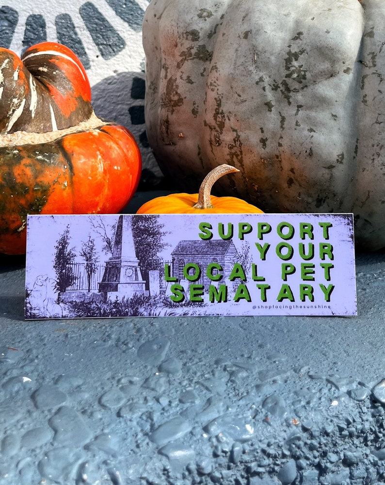 May include: A rectangular sign with the text "SUPPORT YOUR LOCAL PET SEMATARY" in green letters. The sign has a light purple background with a black and white illustration of a cemetery. The sign is in front of pumpkins.