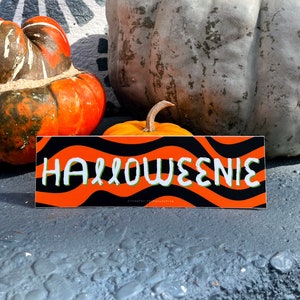 May include: A rectangular sticker with the word "HALLOWEENIE" in white, outlined in green, on an orange and black wavy background. The sticker is displayed in front of pumpkins and gourds, suggesting a Halloween theme.