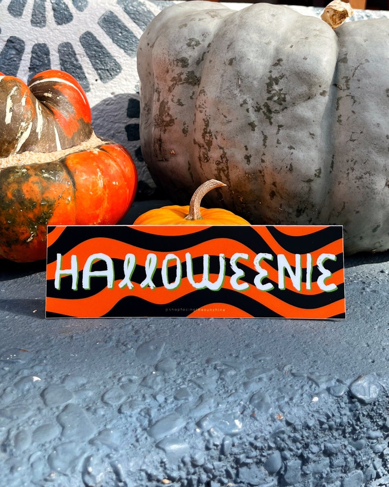 May include: A rectangular sticker with the word "HALLOWEENIE" in white, outlined in green, on an orange and black wavy background. The sticker is displayed in front of pumpkins and gourds, suggesting a Halloween theme.