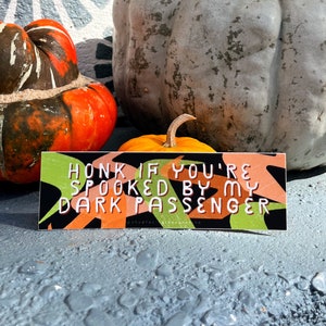 May include: A rectangular sticker with the text "HONK IF YOU'RE SPOOKED BY MY DARK PASSENGER" in white letters. The sticker has a black background with green and orange abstract shapes. The sticker is placed in front of pumpkins.