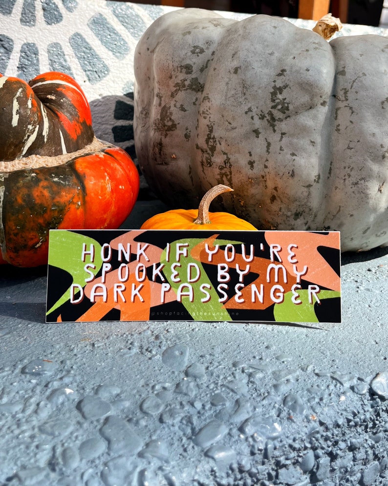 May include: A rectangular sticker with the text "HONK IF YOU'RE SPOOKED BY MY DARK PASSENGER" in white letters. The sticker has a black background with green and orange abstract shapes. The sticker is placed in front of pumpkins.
