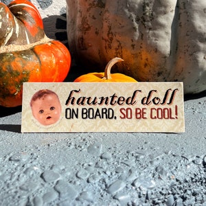 May include: A rectangular sign with the text "haunted doll ON BOARD, SO BE COOL!" features a vintage doll image. The sign is set against a backdrop of pumpkins and gourds, perfect for Halloween.