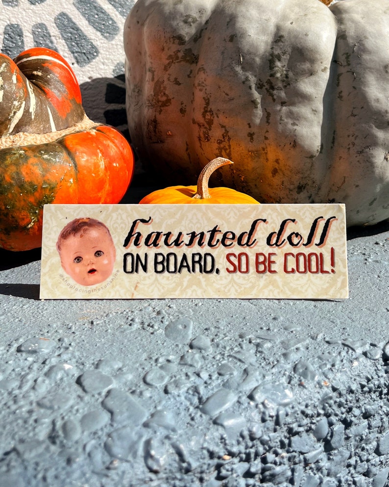 May include: A rectangular sign with the text "haunted doll ON BOARD, SO BE COOL!" features a vintage doll image. The sign is set against a backdrop of pumpkins and gourds, perfect for Halloween.