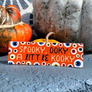 May include: An orange rectangular sign with the text "Spooky, Oooky, A Little Kooky" and various eye illustrations. The sign is placed in front of pumpkins in shades of orange and gray.