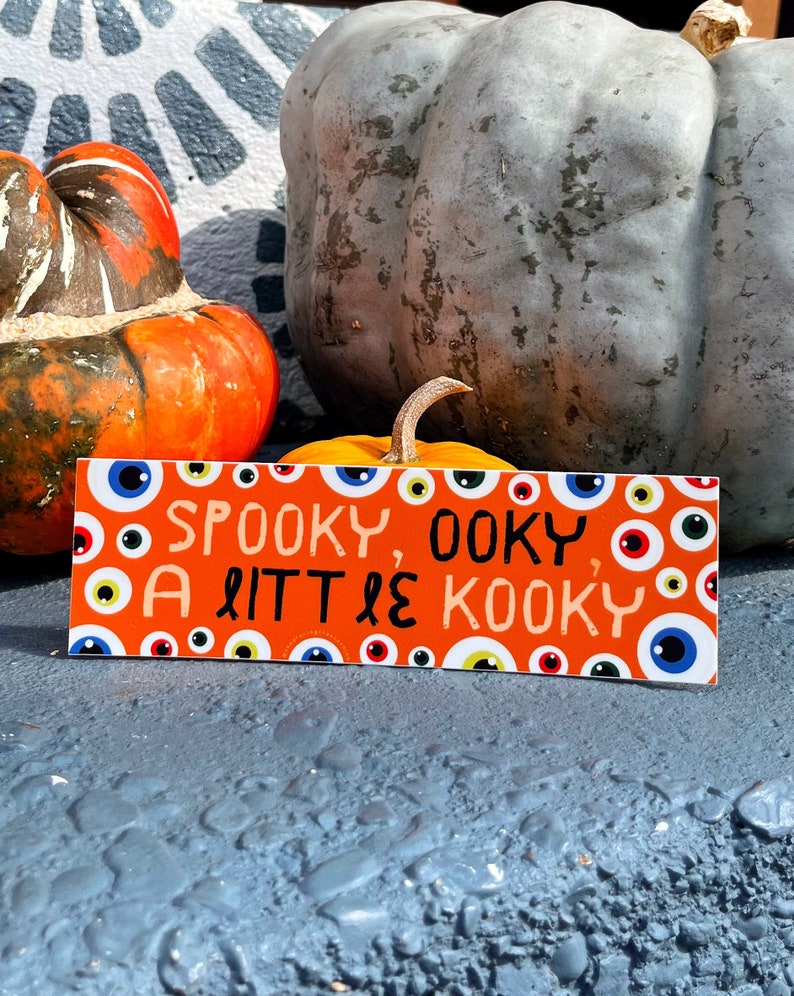 May include: An orange rectangular sign with the text "Spooky, Oooky, A Little Kooky" and various eye illustrations. The sign is placed in front of pumpkins in shades of orange and gray.