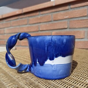 Può includere: Una tazza in ceramica blu con una striscia bianca e un manico attorcigliato. La tazza ha una smaltatura blu e bianca unica.