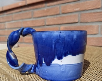 Puede incluir: Una taza de cerámica azul con una franja blanca y un asa retorcida. La taza tiene un esmalte azul y blanco único.