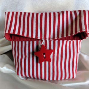 Puede incluir: Un pequeño recipiente de tela con rayas verticales rojas y blancas. Cuenta con un cierre de botón rojo en forma de estrella. El interior y el borde superior doblado son rojos.