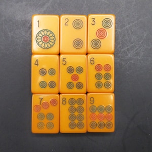 May include: A set of nine rectangular yellow mahjong tiles arranged in a three-by-three grid. Each tile features a black numeral from one to nine in the top left corner and circular floral-patterned icons in red and green ink on the surface.