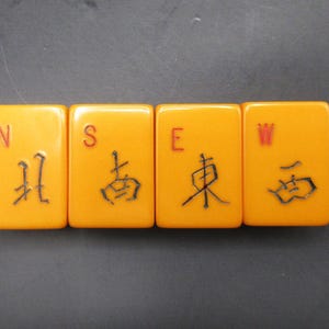 May include: Four rectangular, orange Mahjong tiles arranged horizontally. Each tile displays a direction: North (N), South (S), East (E), and West (W). The directions are written in English and Chinese characters.