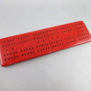 May include: A rectangular red cribbage board featuring a marbled texture and a series of small, evenly spaced holes arranged in rows across the surface. The board is constructed from two connected sections that form a single, flat playing surface.