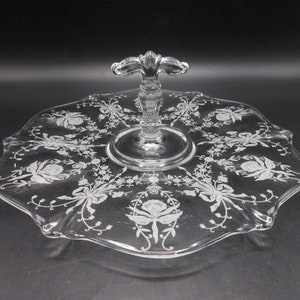 May include: Clear glass serving tray with a floral design. The tray has a scalloped edge and a central handle. The floral pattern is etched into the glass, creating a delicate, elegant look. The tray is suitable for serving cakes, pastries, or other treats.