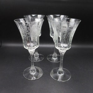 May include: Four clear, etched wine glasses with a floral and cameo design. The glasses have a flared bowl, a long stem, and a round base. The etched design is visible on the bowl of each glass. The glasses are arranged on a black surface.