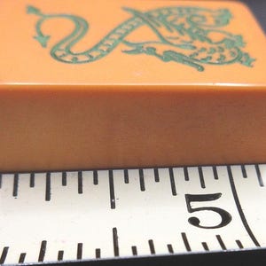 May include: An orange rectangular mahjong tile featuring a green dragon illustration on its top surface, positioned next to a white measuring tape that shows the tile width is approximately 1.25 inches across the scale from 4 to 5 inches.