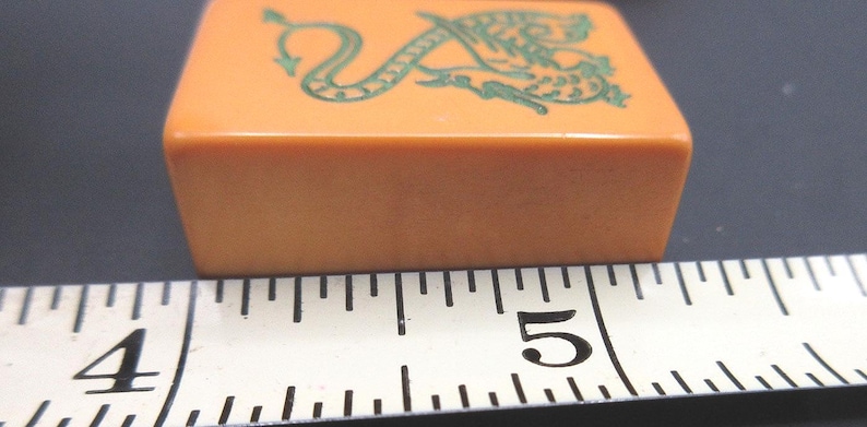 May include: An orange rectangular mahjong tile featuring a green dragon illustration on its top surface, positioned next to a white measuring tape that shows the tile width is approximately 1.25 inches across the scale from 4 to 5 inches.