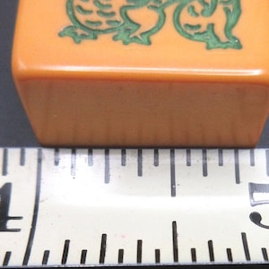 May include: A rectangular orange mahjong tile featuring a green engraved dragon design. The tile is positioned directly above a white measuring tape, which indicates that the width of the tile is approximately 1.25 inches in total length.