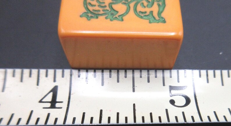 May include: A rectangular orange mahjong tile featuring a green engraved dragon design. The tile is positioned directly above a white measuring tape, which indicates that the width of the tile is approximately 1.25 inches in total length.