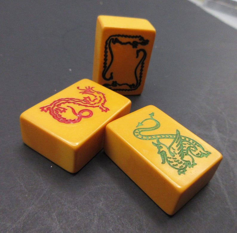 May include: Three rectangular yellow mahjong tiles. One tile features a black engraved border design, one tile displays a red engraved dragon illustration, and the third tile shows a green engraved dragon illustration with wings and a tail.