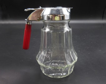 May include: A clear glass syrup dispenser with a red handle and a silver top. The dispenser has a textured, fluted design.