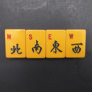 May include: Four rectangular yellow mahjong tiles arranged in a horizontal row. Each tile features a red letter N, S, E, or W in the top left corner and a corresponding black Chinese character representing North, South, East, and West below it.