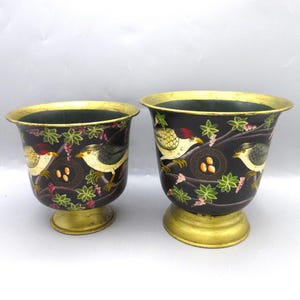May include: Two decorative black metal planters with gold rims and bases. Each planter features a painted design of birds, nests, and floral elements. The planters are of different sizes and are suitable for indoor or outdoor use.