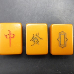 May include: Three rectangular Mahjong tiles with orange-yellow surfaces. Each tile features a different symbol: a red Chinese character, a black Chinese character, and a black geometric design. The tiles are arranged side by side.