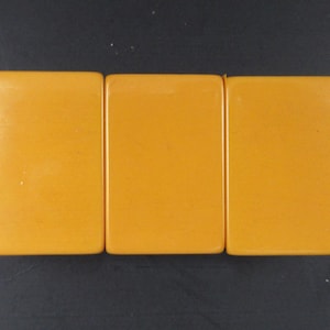 May include: Three rectangular mahjong tiles with a smooth, solid mustard yellow finish. Each tile features rounded corners and a flat, unadorned surface. The tiles are arranged side-by-side in a horizontal row, displaying a uniform color and texture.