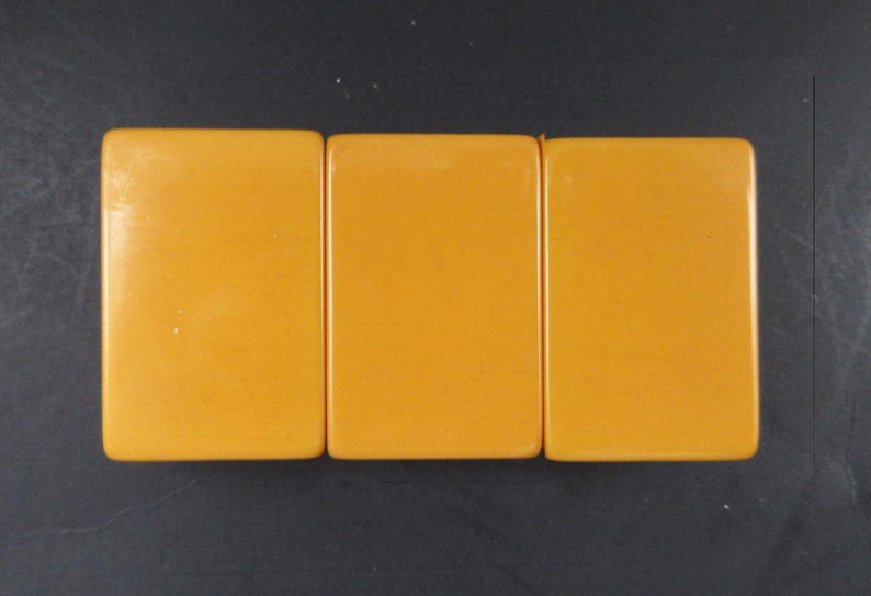 May include: Three rectangular mahjong tiles with a smooth, solid mustard yellow finish. Each tile features rounded corners and a flat, unadorned surface. The tiles are arranged side-by-side in a horizontal row, displaying a uniform color and texture.