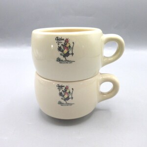 May include: Two cream-colored ceramic mugs stacked on top of each other. Each mug has a rounded shape with a handle and a graphic of a rooster holding a fishing rod. The text "Chicken of the Rough" is above the rooster.